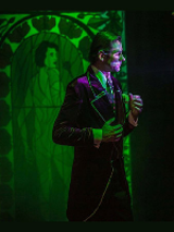 Zdjęcie z przedstawienia - mężczyzna stoj ący przed lustrem, w tle obraz-witraż w zielonych kolorach przedstawiający kobietę.