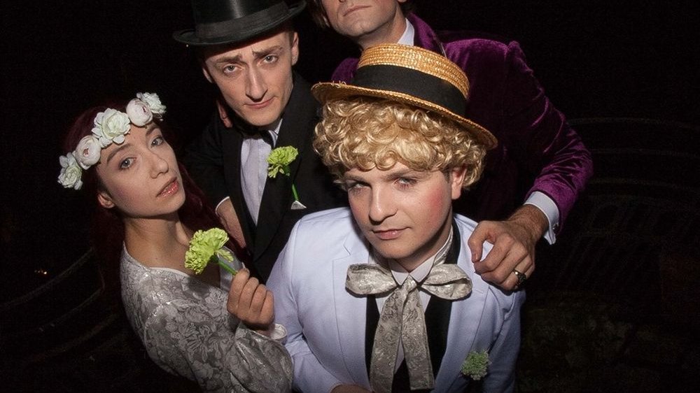 Portret grupy aktorów w kostiumach teatralnych. Mężczyźni mają na sobie garnitury i kapelusze, kobieta ślubną sukienkę i wianek.