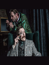 Zdjęcie z przedstawienia - jeden mężczyzna siedzący w fotelu z lupą przy oku, za nim stoi drugi mężczyna z fajką.