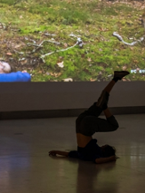 Zdjęcie ze spektaklu - tancerka leżąca na podłodze z uniesionymi pionowo nogami. W tle fragment zdjęcia wyświetlonego na ścianie, przedstawiającego osobę w podobnej pozie na trawie przy drzewie.