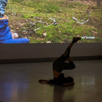 Zdjęcie ze spektaklu - tancerka leżąca na podłodze z uniesionymi pionowo nogami. W tle fragment zdjęcia wyświetlonego na ścianie, przedstawiającego osobę w podobnej pozie na trawie przy drzewie.