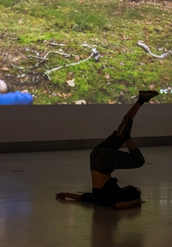 Zdjęcie ze spektaklu - tancerka leżąca na podłodze z uniesionymi pionowo nogami. W tle fragment zdjęcia wyświetlonego na ścianie, przedstawiającego osobę w podobnej pozie na trawie przy drzewie.