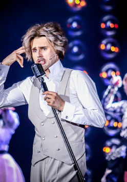 Zdjęcie ze spektaklu - mężczyzna w bialej koszuli i szarych kamizelce i spodniach śpiewający do mikrofonu. W tle dwie aktorki przy mikrofonach.