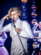 Zdjęcie ze spektaklu - mężczyzna w bialej koszuli i szarych kamizelce i spodniach śpiewający do mikrofonu. W tle dwie aktorki przy mikrofonach.