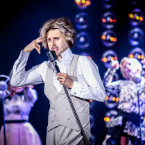 Zdjęcie ze spektaklu - mężczyzna w bialej koszuli i szarych kamizelce i spodniach śpiewający do mikrofonu. W tle dwie aktorki przy mikrofonach.