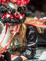 Zdjęcie ze spektaklu - kobieta z czepcem w biało-czerwono-białych kolorach na głowie, leżąca i patrzaca na mężczyznę, którego twarz jest niewidoczna.