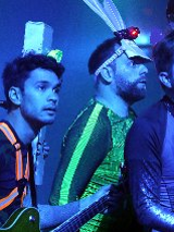 Czwórka artystów w kolorowych, kostiumach scenicznych występuje na estradzie w niebieskim oświetleniu. Muzycy trzymają instrumenty, w tym gitarę oraz saksofon, i patrzą z przejęciem w jednym kierunku. Całość ma dynamiczny, teatralny charakter z wykorzystaniem nietypowych rekwizytów na głowach wykonawców.