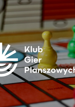 Pionki i kostki do gry planszowej rozstawione na planszy, na nich napis Klub Gier Planszowych i logo przypominające słońce