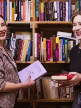 Dwie uśmiechnięte kobiety stoją przed bibliotecznym regałem, w rękach trzymają książki.