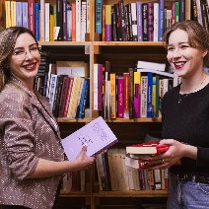 Dwie uśmiechnięte kobiety stoją przed bibliotecznym regałem, w rękach trzymają książki.