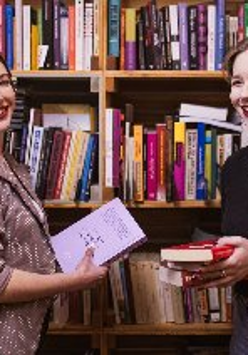 Dwie uśmiechnięte kobiety stoją przed bibliotecznym regałem, w rękach trzymają książki.