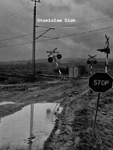 Informacje o wystawie fotografii Stanisława Cioka, czarno-białe zdjęcie zaniedbanego przejazdu kolejowego w pagórkowatym krajobrazie.