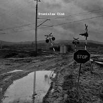 Informacje o wystawie fotografii Stanisława Cioka, czarno-białe zdjęcie zaniedbanego przejazdu kolejowego w pagórkowatym krajobrazie.