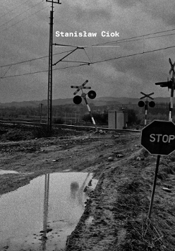 Informacje o wystawie fotografii Stanisława Cioka, czarno-białe zdjęcie zaniedbanego przejazdu kolejowego w pagórkowatym krajobrazie.
