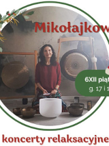 Zdjęcie przedstawia kobietę siedzącą przed dużym gongiem oraz kilkoma misami do śpiewu. Kobieta ma na sobie czerwoną koszulkę. Gong jest zawieszony na suficie za kobietą. Misy do śpiewu są ułożone w półkolu przed nią. Tło stanowi biała ściana. Kobieta ma ciemne włosy i patrzy w obiektyw aparatu. Kobieta lekko się uśmiecha.