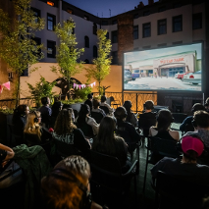 Grupa ludzi siedzi na krzesłach i ogląda film na ekranie plenerowym. Widzowie mają na uszach słuchawki, przez które słyszą dźwięk filmu. Na balkonie, na którym siedzą postawiono małe drzewka, a między nimi rozwieszono różowe girlandy.
