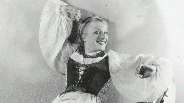 Czarno-białe zdjęcie uśmiechniętej kobiety w ludowym stroju, z podniesioną nogą i ręką.