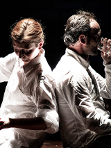 Kobieta i mężczyzna, siedzą obróceni do siebie plecami. Kobieta nalewa z czajnika wodę do szklanki, mężczyzna pije wodę ze szklanki.