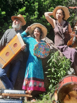 Mężczyzna i kobiety w średnim wieku ubrani w kolorowe stroje. Na głowach mają słomkowe kapelusze a w rękach trzymają różne instrumenty muzyczne. Przed nimi leżą inne instrmenty. Za nimi drzewa i kolorowe kwiaty.