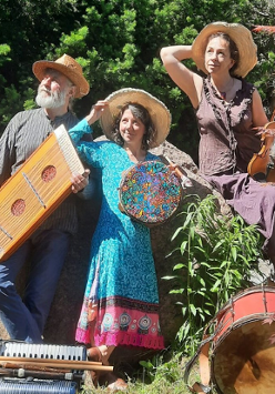 Mężczyzna i kobiety w średnim wieku ubrani w kolorowe stroje. Na głowach mają słomkowe kapelusze a w rękach trzymają różne instrumenty muzyczne. Przed nimi leżą inne instrmenty. Za nimi drzewa i kolorowe kwiaty.