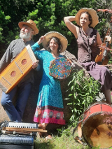 Mężczyzna i kobiety w średnim wieku ubrani w kolorowe stroje. Na głowach mają słomkowe kapelusze a w rękach trzymają różne instrumenty muzyczne. Przed nimi leżą inne instrmenty. Za nimi drzewa i kolorowe kwiaty.