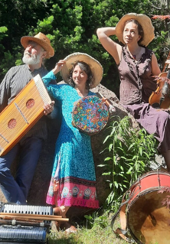 Mężczyzna i kobiety w średnim wieku ubrani w kolorowe stroje. Na głowach mają słomkowe kapelusze a w rękach trzymają różne instrumenty muzyczne. Przed nimi leżą inne instrmenty. Za nimi drzewa i kolorowe kwiaty.