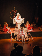 Grupa osób w słowiańskich strojach z wzorami ludowymi siedzi na drewnianej scenie teatralnej.