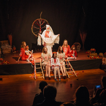 Grupa osób w słowiańskich strojach z wzorami ludowymi siedzi na drewnianej scenie teatralnej.