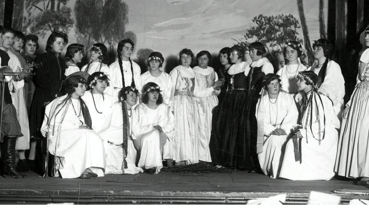 Teatralna grupa młodych osób stojących w rzędzie w starodawnych strojach. W tle teatralna dekoracja.