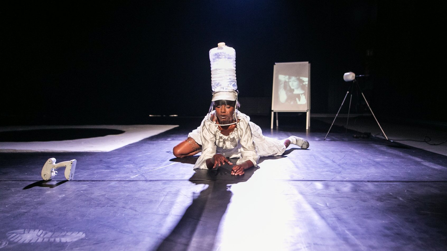 Performerka w białym stroju i plastikowej butli na głowie czołga się w stronę obiektywu.
