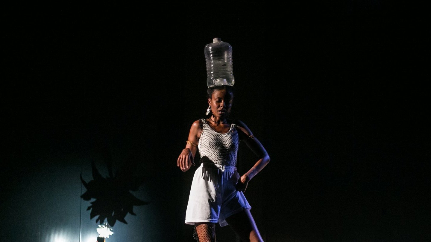 Kobieta w białym stroju i z plastikową butlą na głowie tańczy wśród rozsypanego cukru. Stoi na zgiętych nogach i wystawia jedną rękę w stronę obiektywu.