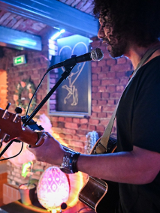 Mężczyzna z kręconymi włosami. Trzyma w rękach gitarę. Śpiewa do mikrofonu. W tle ceglana ściana z ozdobami.