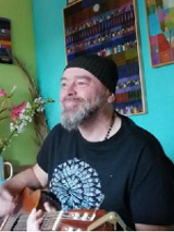 Tomasz Budzyński w czarnej czapce i ciemnej koszulce z gitarą w mieszkaniu. Po lewej zielona ściana, po prawej turkusowa. Na ścianie obrazy.