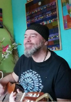 Tomasz Budzyński w czarnej czapce i ciemnej koszulce z gitarą w mieszkaniu. Po lewej zielona ściana, po prawej turkusowa. Na ścianie obrazy.