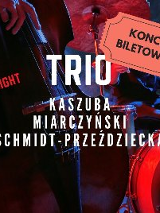 Zdjęcie przedstawia plakat koncertu jazzowego. Przedstawia trio muzyków występujących na scenie. Muzycy grają na kontrabasie, perkusji i fortepianie. Plakat jest czerwono-czarny i zawiera duży bilet z napisem "KONCERT BILETOWAN". Plakat zawiera również nazwiska muzyków: Kaszuba, Miarczyński i Schmidt-Przeździecka. Całość oddaje dynamiczną i żywą atmosferę występu jazzowego na żywo.