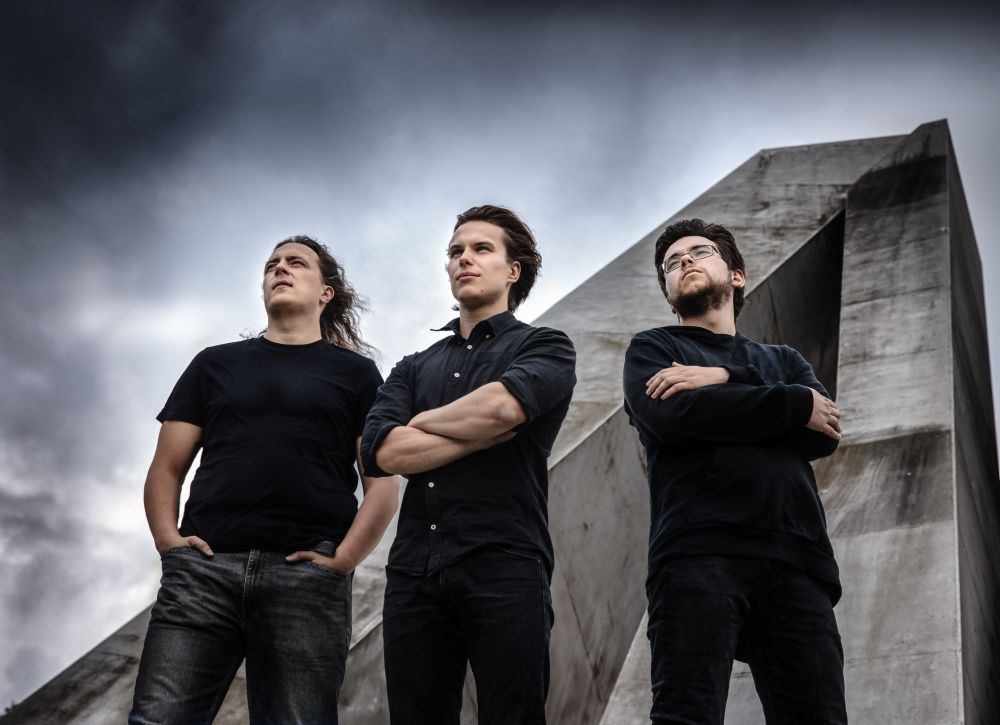 Trzech młodych mężczyzn stoi na tle zachmurzonego nieba i betonowego pomnika.