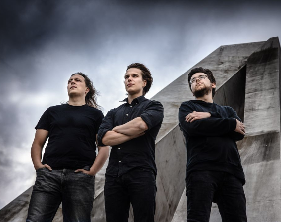 Trzech młodych mężczyzn stoi na tle zachmurzonego nieba i betonowego pomnika.