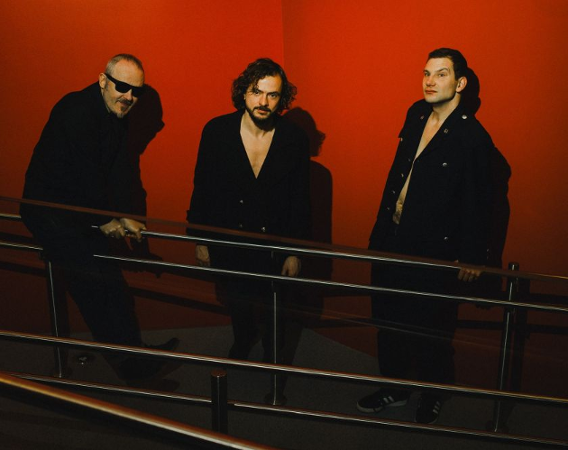 Trzech muzyków pozuje na tle czerwonej ściany.