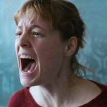 Zbliżenie na twarz krzyczącej kobiety. Kobieta ma rude włosy i czerwoną bluzkę. Stoi na tle tablicy w klasie.