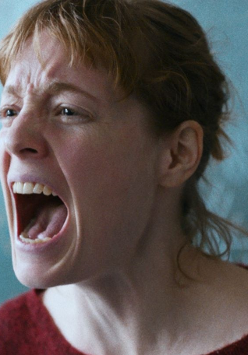 Zbliżenie na twarz krzyczącej kobiety. Kobieta ma rude włosy i czerwoną bluzkę. Stoi na tle tablicy w klasie.