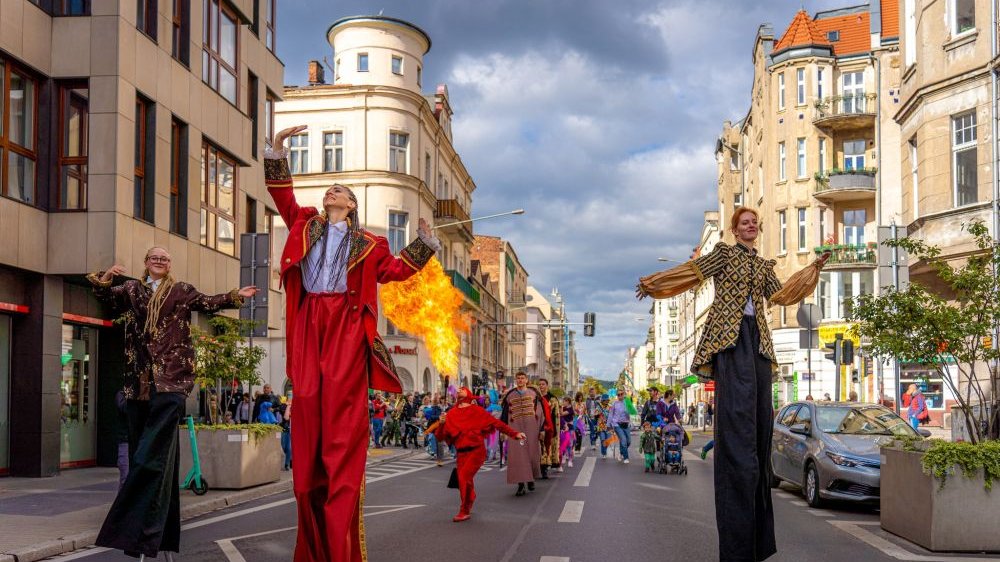 Trzy kobiety idące na szczudłach prowadzą kolorowy przemarsz ulicą miasta.