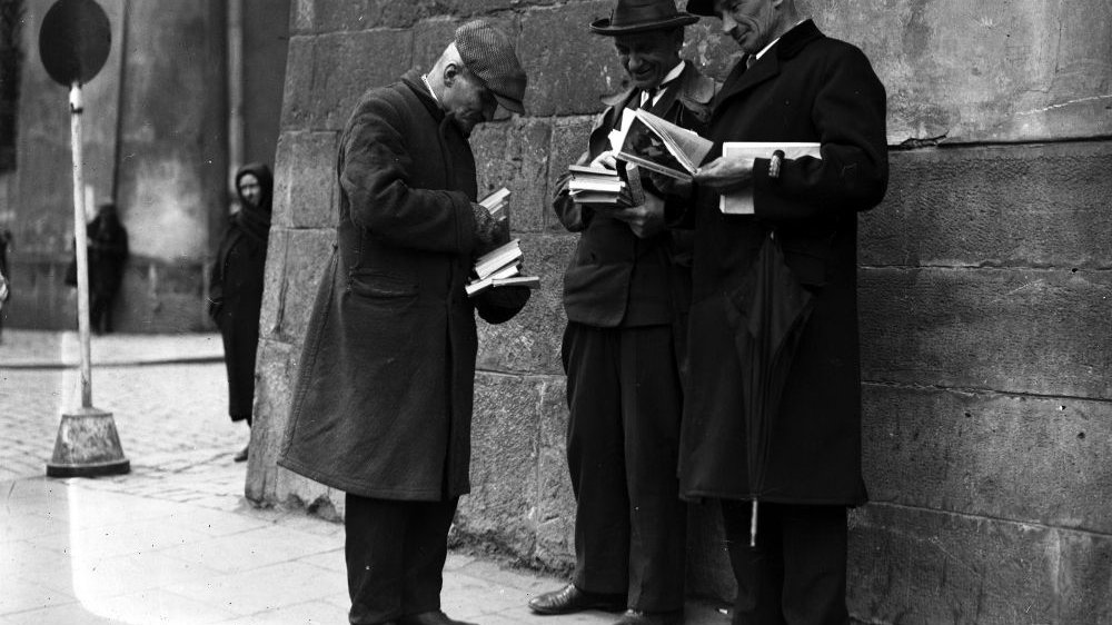 Czarno-białe zdjęcie trzech mężczyn w płaszczach i nakryciach głowy. Stoją na ulicy, w rękach trzymają książki.