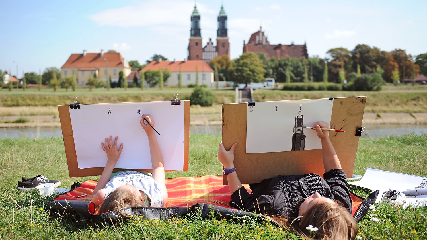 Mama z dzieckiem leżą na kocu na trawie i rysują Katedrę Poznańską widzianą z drugiego brzegu rzeki Warty