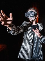 Na zdjęciu stoi kobieta w garniturze w paski. Jej ramię wyciąga się w stronę publiczności. Za nią znajduje się czerwona scena za kulisami.