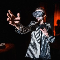 Na zdjęciu stoi kobieta w garniturze w paski. Jej ramię wyciąga się w stronę publiczności. Za nią znajduje się czerwona scena za kulisami.