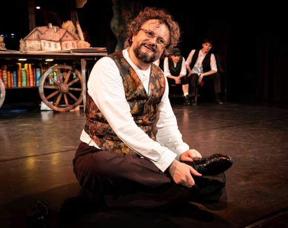 Uśmiechnięty mężczyzna w złotej kamizelce i białej koszuli siedzi na scenie.