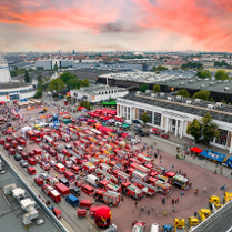 Widok z góry na Międzynarodowe Targi Poznańskie. Na placu głównym stoją zaparkowane wozy strażackie w kolorze czerwonym. Wokół nich gromadzą się ludzie, którzy oglądają pojazdy i robią zdjęcia. W oddali rozpościera się panorama Poznania.