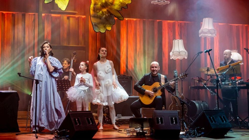 Artystka w długiej, fioletowej sukni, śpiewa obok dwóch dziewczynek ubranych w baletowe sukienki. Na scenie siedzi także gitarzysta. Wszystko jest oświetlone pomarańczowym światłem.