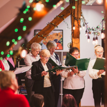 Grupa śpiewającycj osób w udekorowanym świątecznie pomieszczeniu.