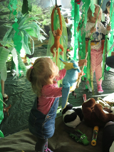 Mała dziewczynka bawi się porozwieszanymi zabawkami. Pomieszczenie przypomina dżunglę, w której wiszą liany i zwierzęta.